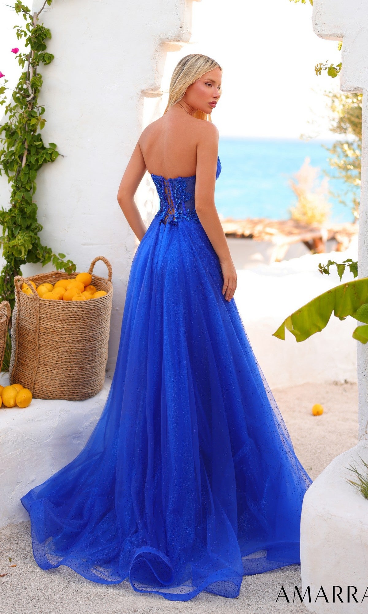 A woman wears the Amarra 89488 long prom dress, which features a strapless embellished corset bodice and glitter tulle A-line skirt, standing outdoors near white walls with baskets of lemons and the sea in view.